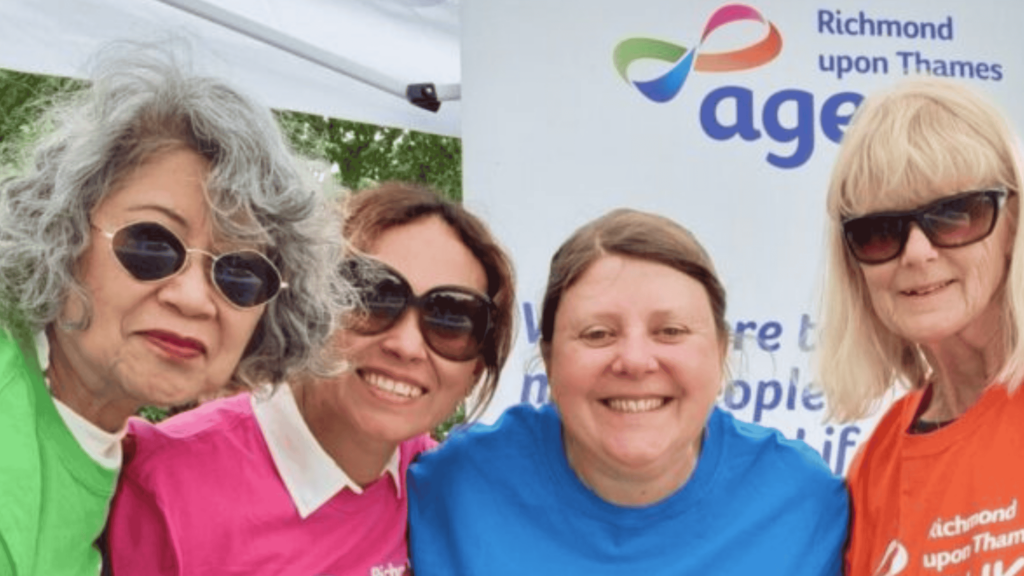 A group of Age UK Volunteers in different coloured t-shirts smile into the camera, standing in front of an Age UK banner.