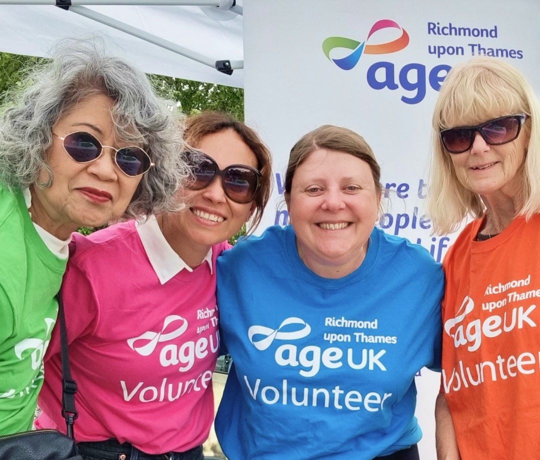 A group of Age UK Volunteers in different coloured t-shirts smile into the camera, standing in front of an Age UK banner.