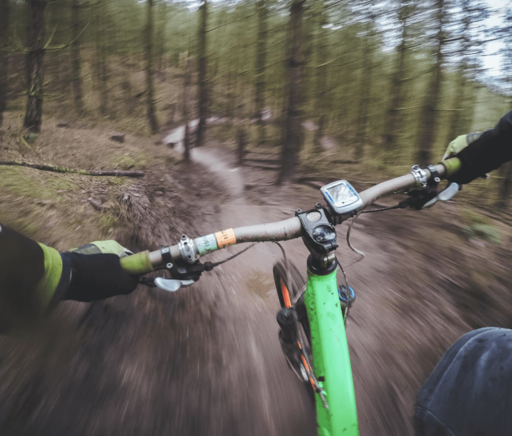 A dynamic action shot of a cyclist navigating a narrow, winding trail in a forest. The image is taken from the cyclist's perspective, focusing on the handlebars of a green mountain bike. The forest is dense with tall trees, and the path appears muddy and challenging, adding to the sense of speed and adventure.